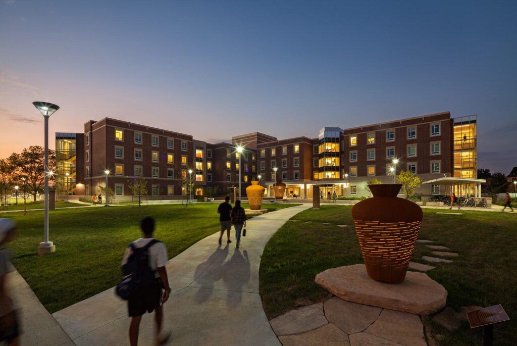 A modern college residence with students walking along a path, surrounded by contemporary sculptures and warm evening lighting.