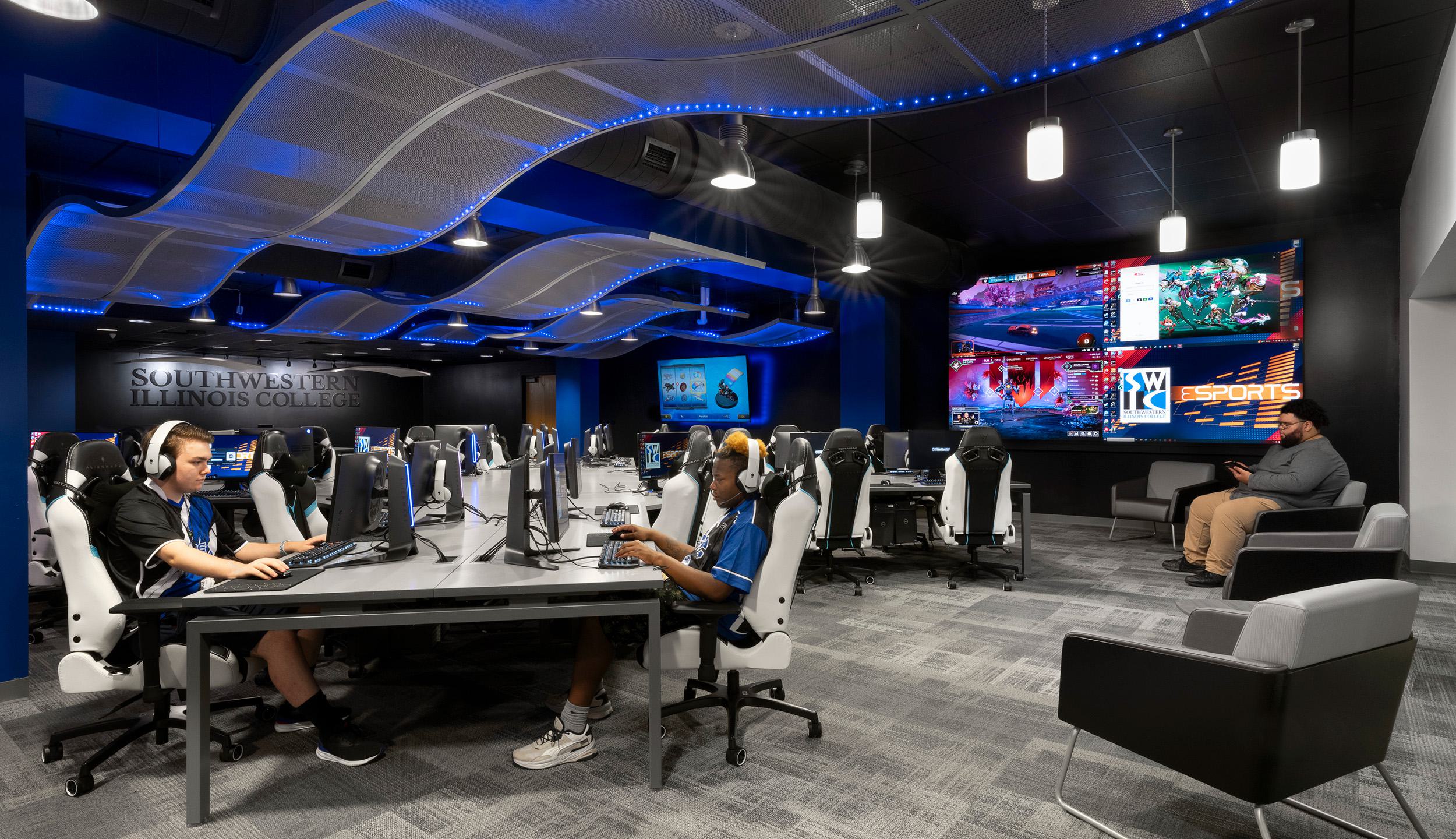 Southwestern Illinois College Esports arena with two students on computers in the foreground and another student in the background; large screen in back, blue LED lighting and modern computer station setup.