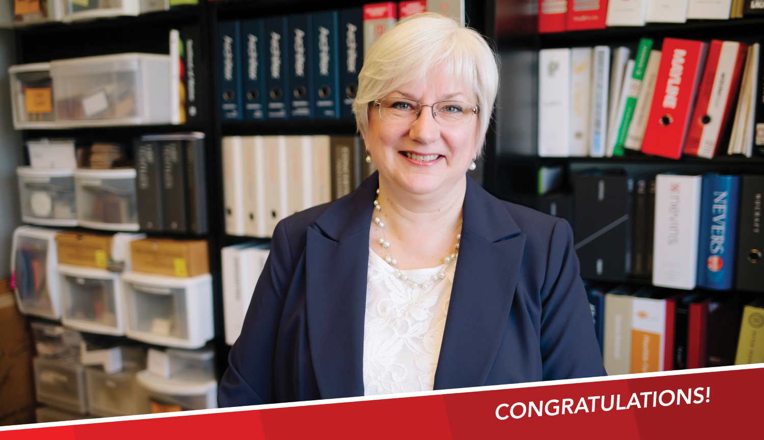 Diane Gilmartin, FGMA CFO, smiling with glasses and short, light hair poses in front of shelves filled with books and binders, with a banner in the corner that reads "Congratulations!"