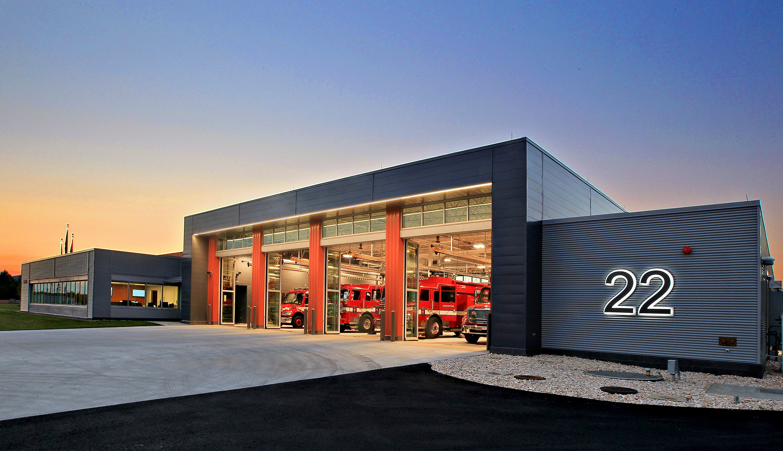 A modern fire station features spacious bays for fire trucks, illuminated signage, and a sleek architectural design against a sunset backdrop.