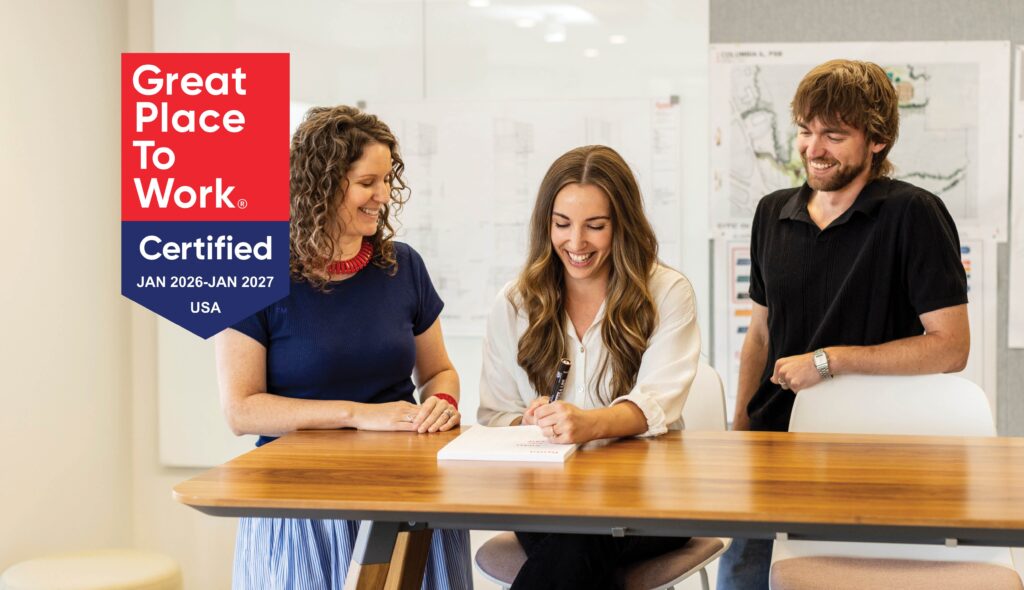 Three colleagues collaborate at a wooden table, sharing smiles and ideas, embodying a friendly and productive work environment.