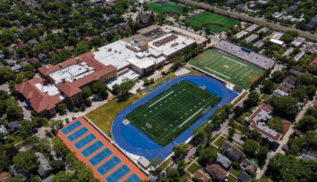 Aerial view of a sports complex featuring a track, football field, tennis courts, and surrounding buildings in a suburban area.