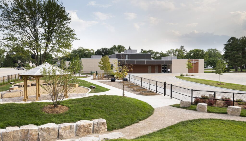 Green grass, trees and a natural playground area features pathways and a fenced-off parking lot with the entry to the sustainable and award-winning Prairie Trails School in the background.