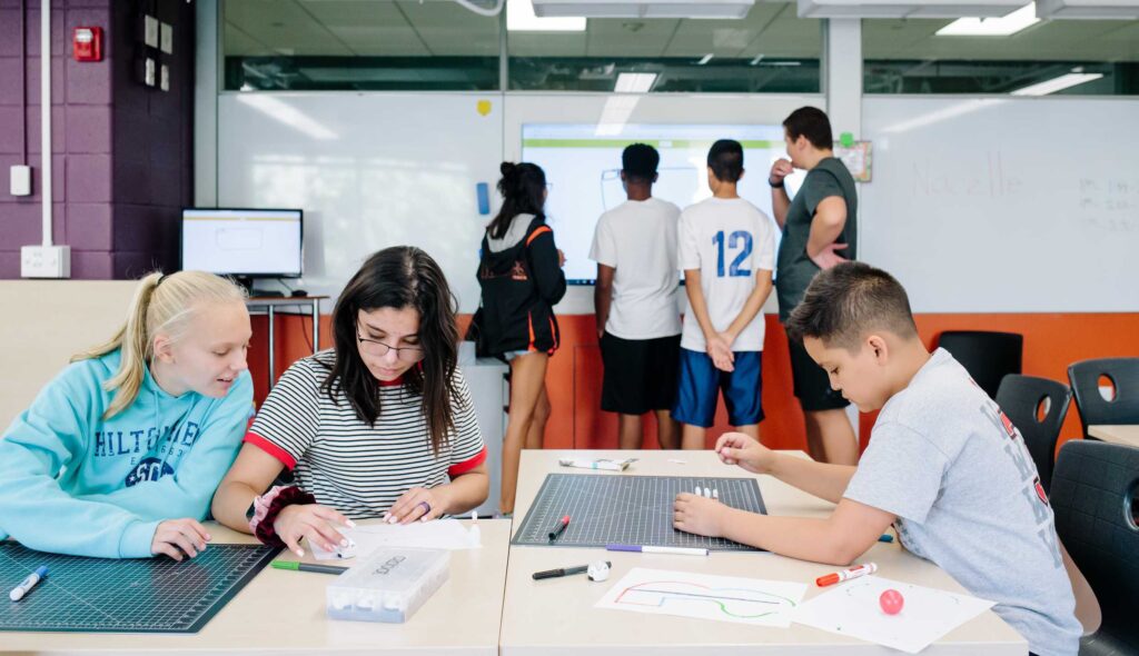 Students collaborate at desks while others observe a presentation on a screen, fostering engagement and teamwork in an educational setting.
