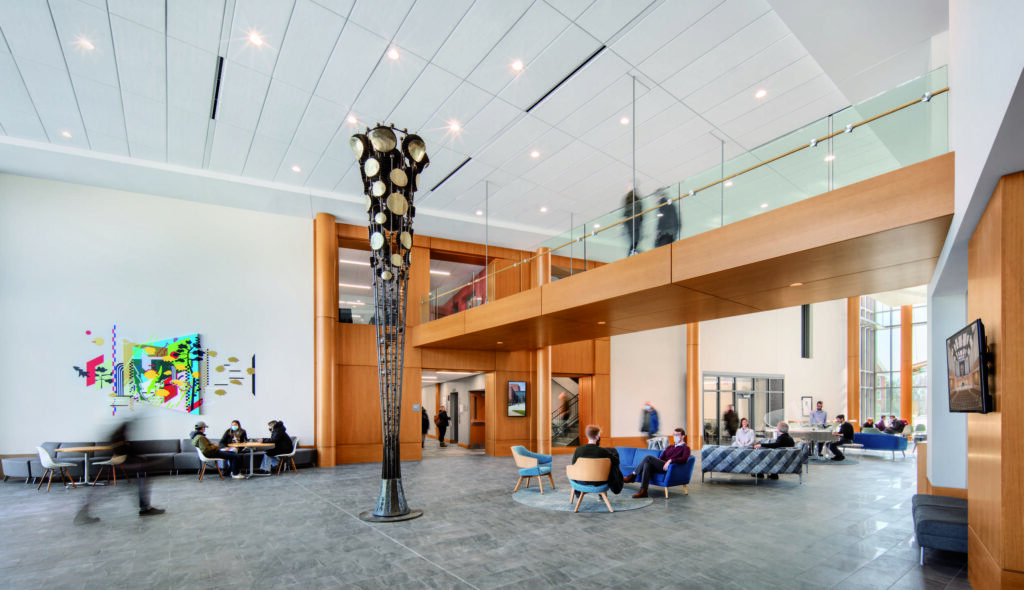 A modern lobby features sleek wooden accents, flexible seating arrangements and people engaging in conversations, with a stylish staircase above.