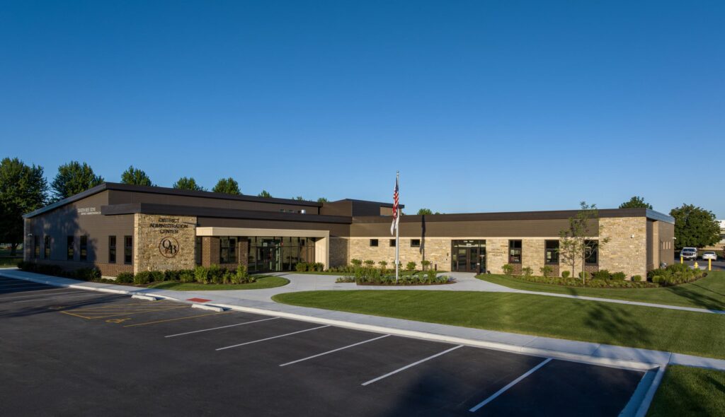 A modern stone and metal building with an American flag, surrounded by a landscaped area and a parking lot under a clear blue sky.