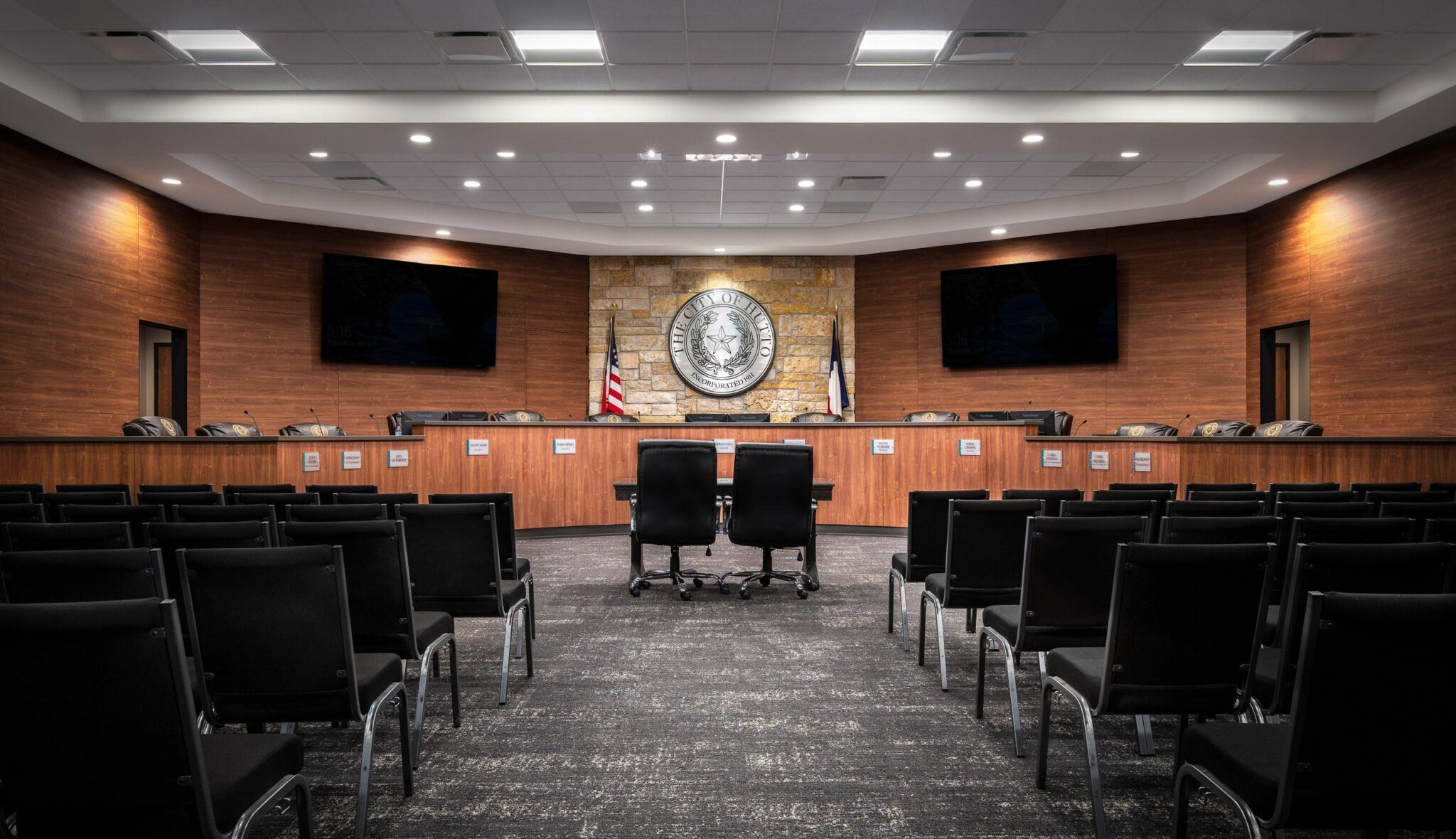 A modern courtroom features a wooden panel backdrop, flags, two chairs at a central desk, and rows of black chairs for spectators.
