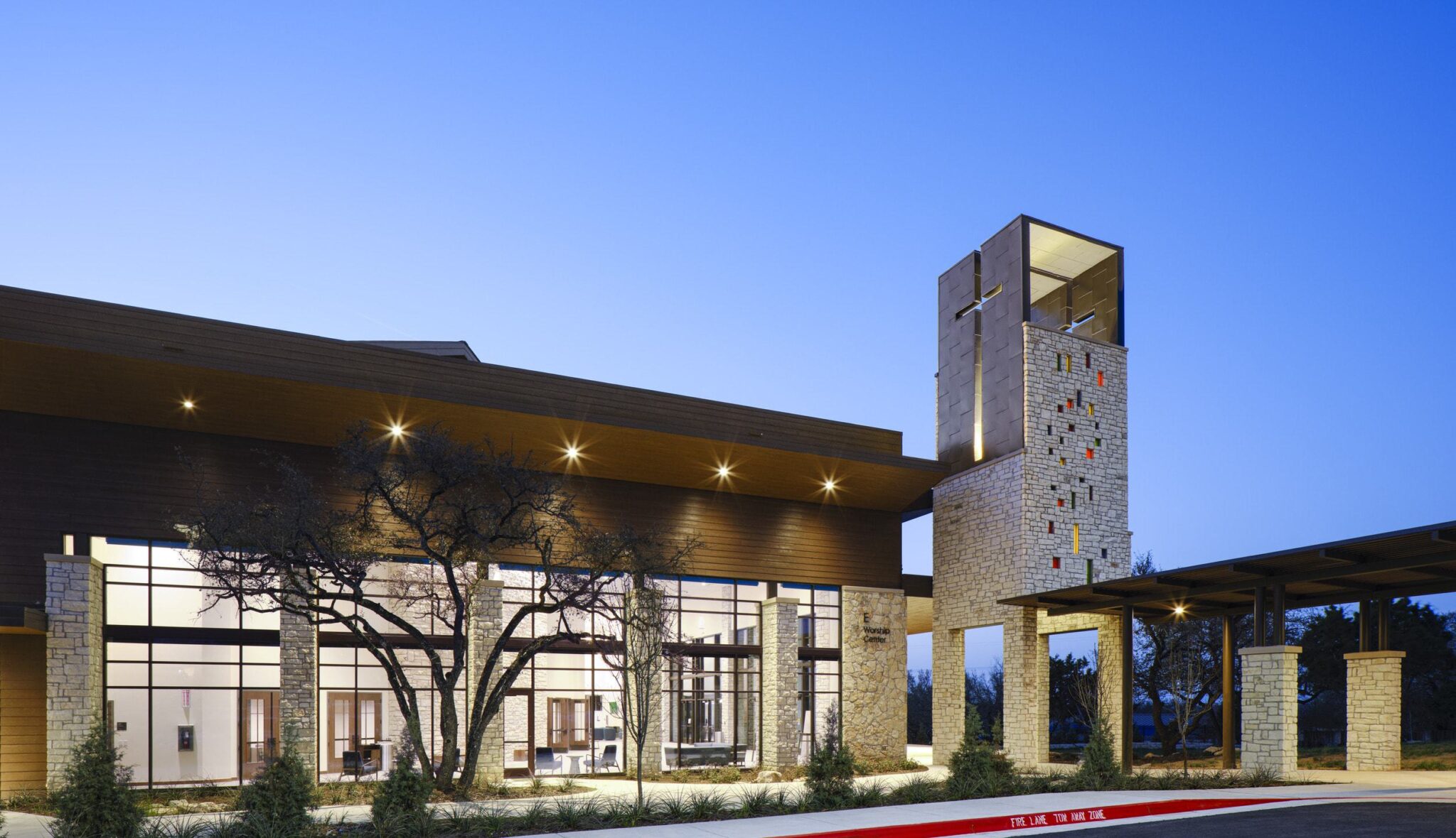 A modern building with large glass windows, featuring a tall stone tower adorned with colorful designs, set against a twilight sky.