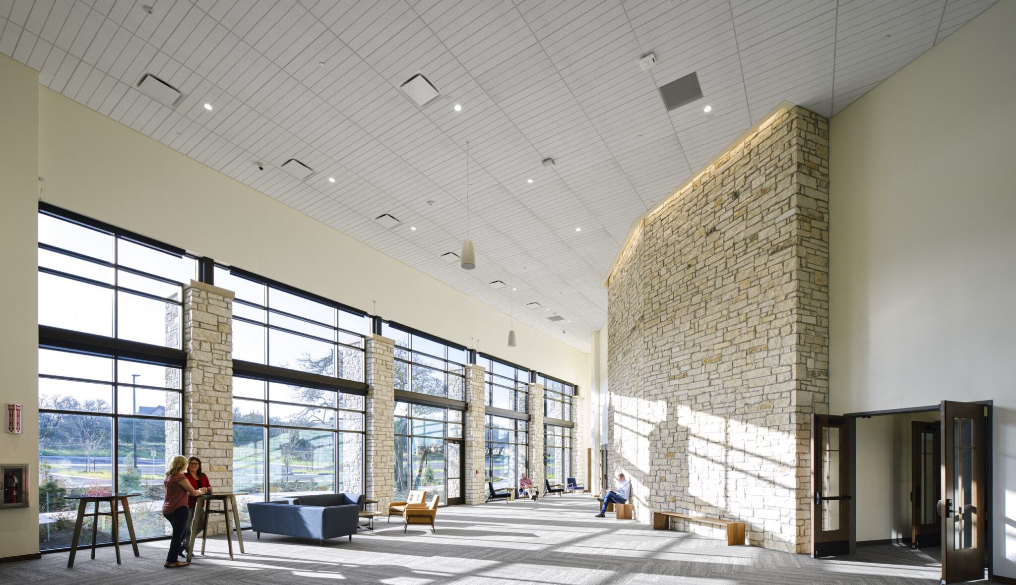 A spacious, modern interior features large windows, a stone wall, and people relaxing in a well-lit, open environment.