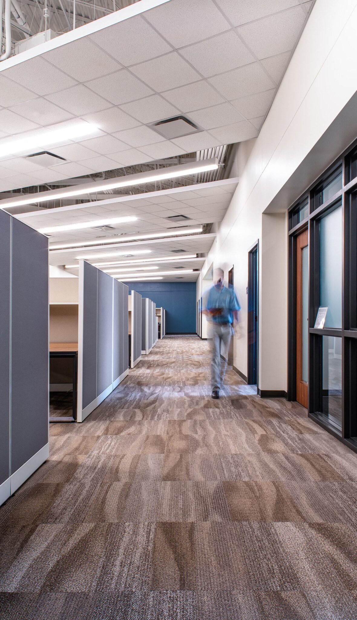 A modern office corridor lined with cubicles, featuring neutral tones and a blurred figure walking towards the camera.