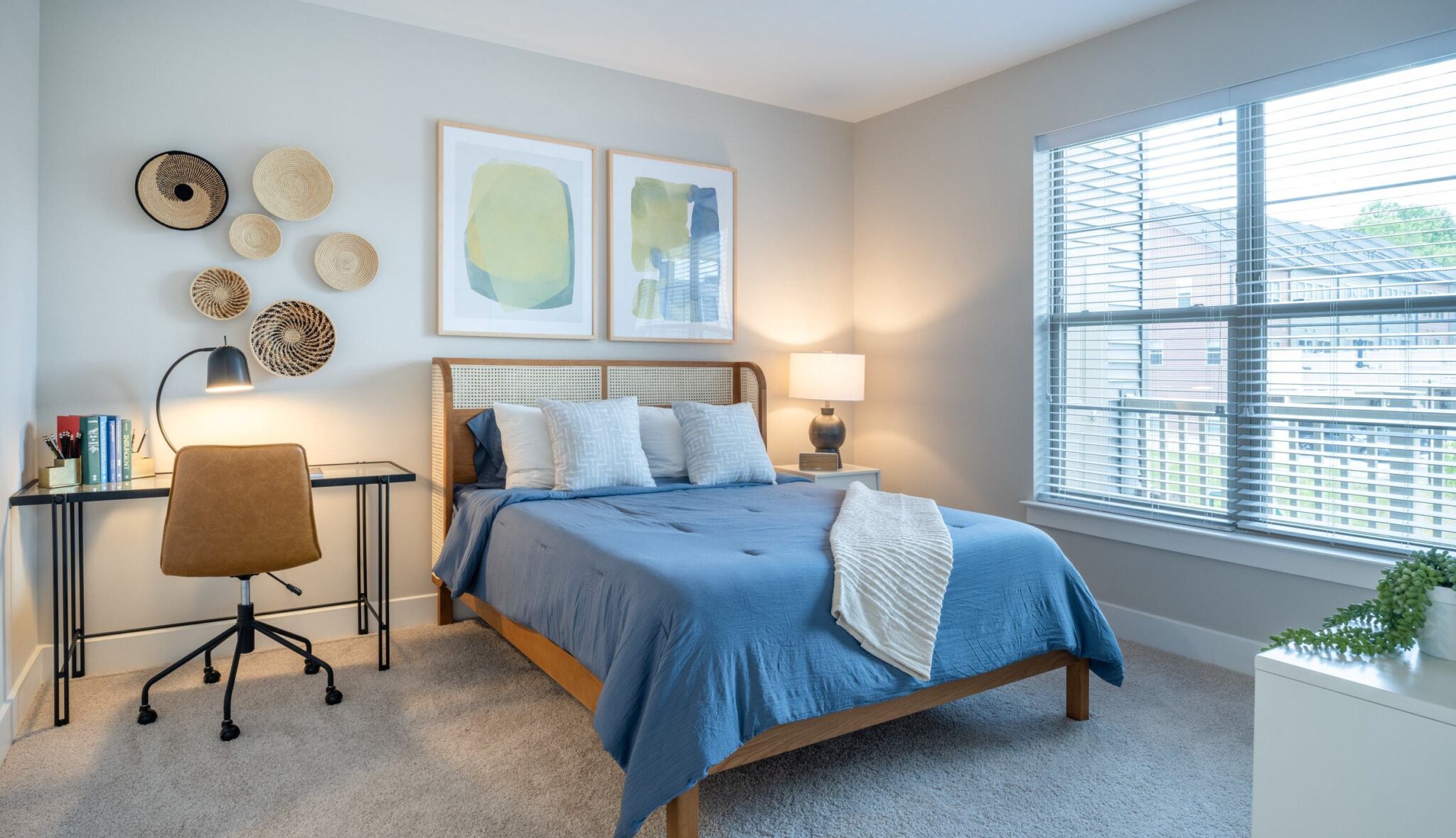A cozy bedroom features a blue bedspread, modern art, a lamp, a desk, and large windows allowing natural light to brighten the space.