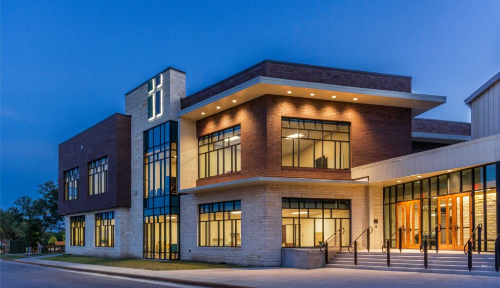 A modern church building features large windows, brick and stone accents, and illuminated entrances, set against a twilight sky.