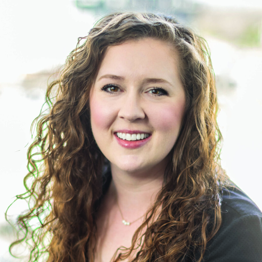A smiling woman with long, curly hair and a cheerful expression, set against a soft, blurred background.