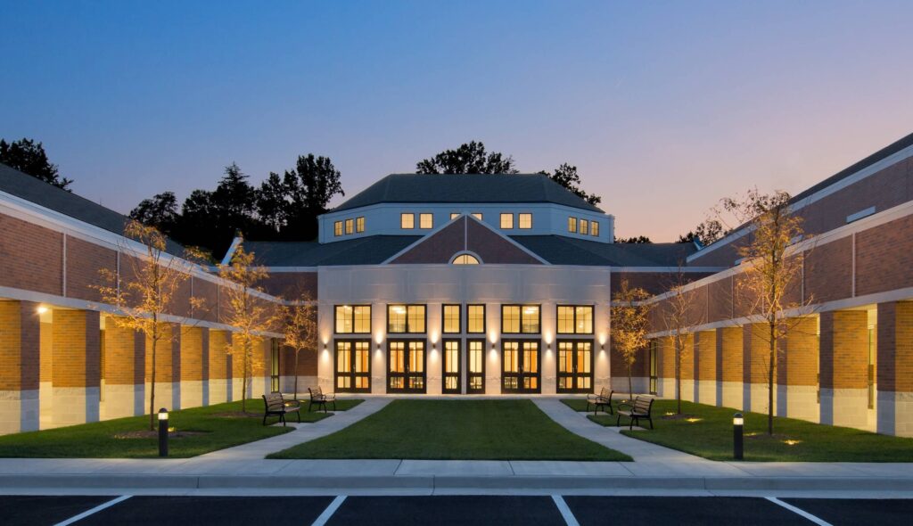 A modern church building featuring brick, large glass doors, surrounded by landscaped greenery and evening twilight, creating a welcoming ambiance.