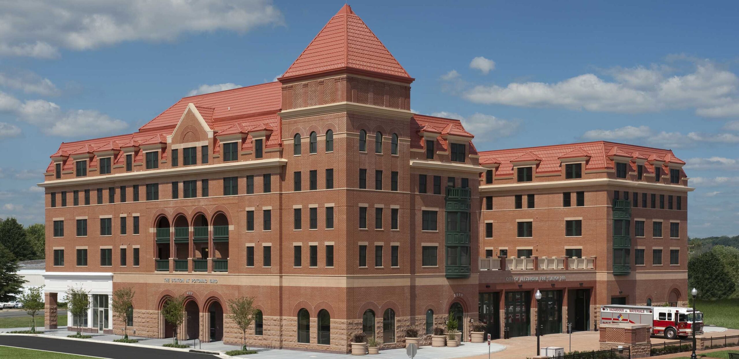 A large, modern brick building features a striking red tiled roof, arched windows, and a welcoming entrance, set against a blue sky.