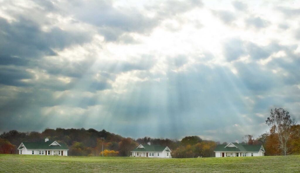 A tranquil landscape features several church cabins on a grassy field, illuminated by soft rays of sunlight breaking through cloudy skies.