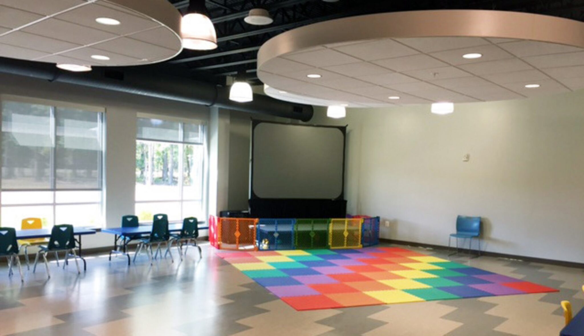 A bright children's playroom with colorful flooring, a play area enclosed by a fence, tables and chairs, and large windows allowing natural light.