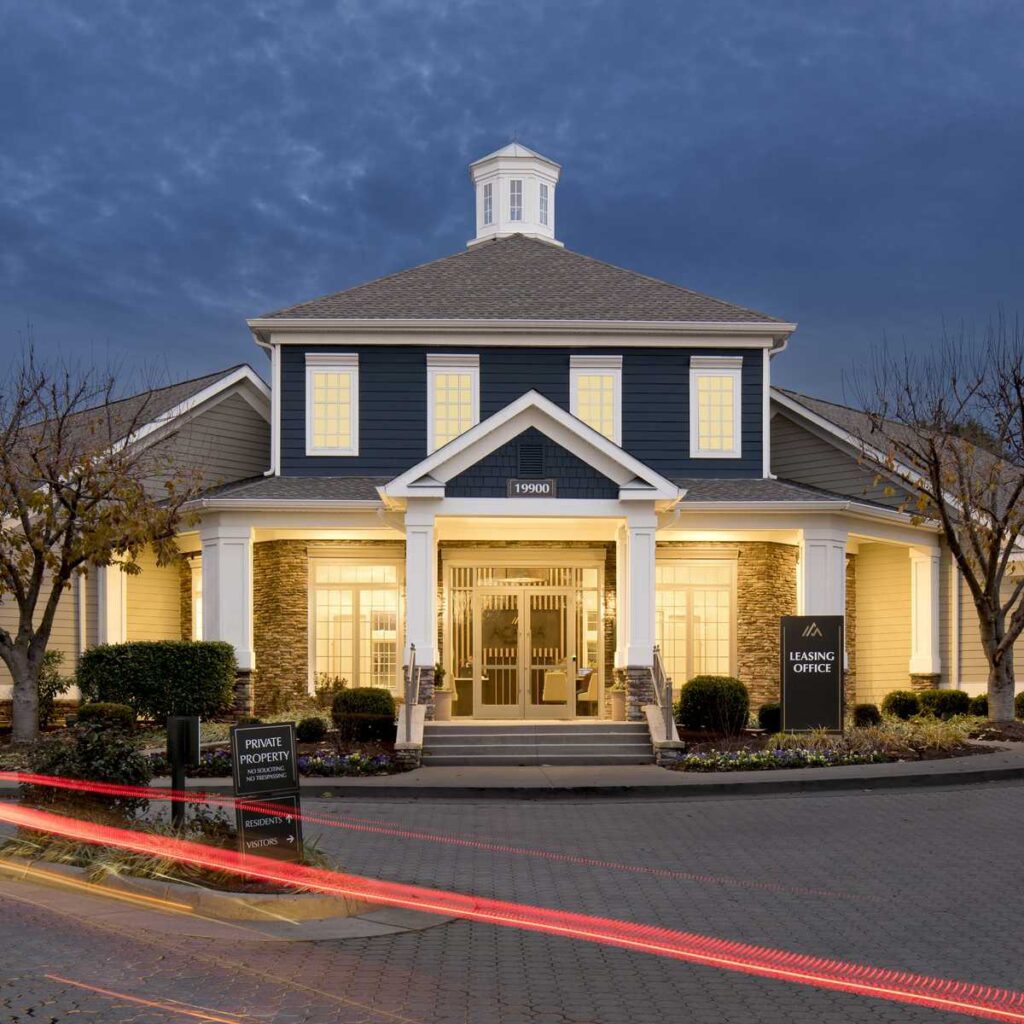 A charming two-story building with a peaked roof, warmly lit windows, and landscaped entry, featuring a leasing office sign out front.
