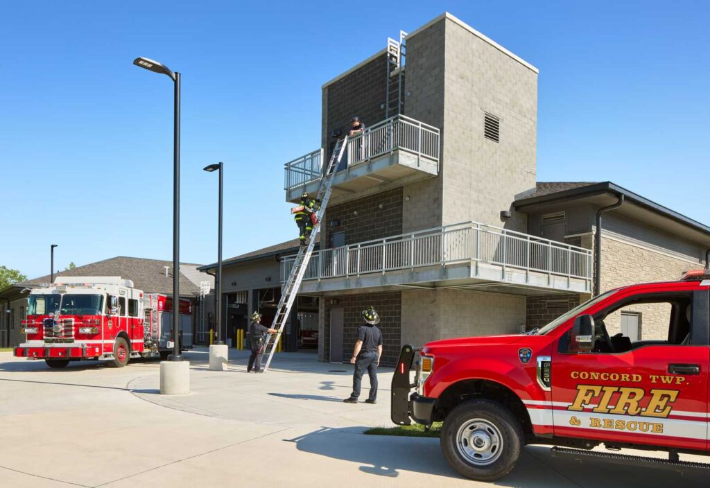 Firefighters training on a building with ladders, surrounded by fire trucks parked at a station in a clear, sunny environment.