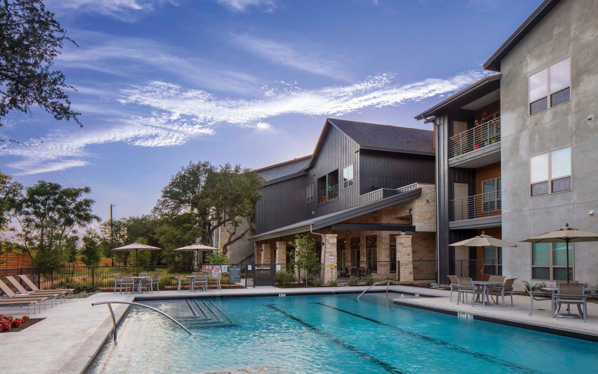 A modern pool area with lounge chairs and umbrellas, surrounded by lush greenery and stylish apartments under a scenic sky.