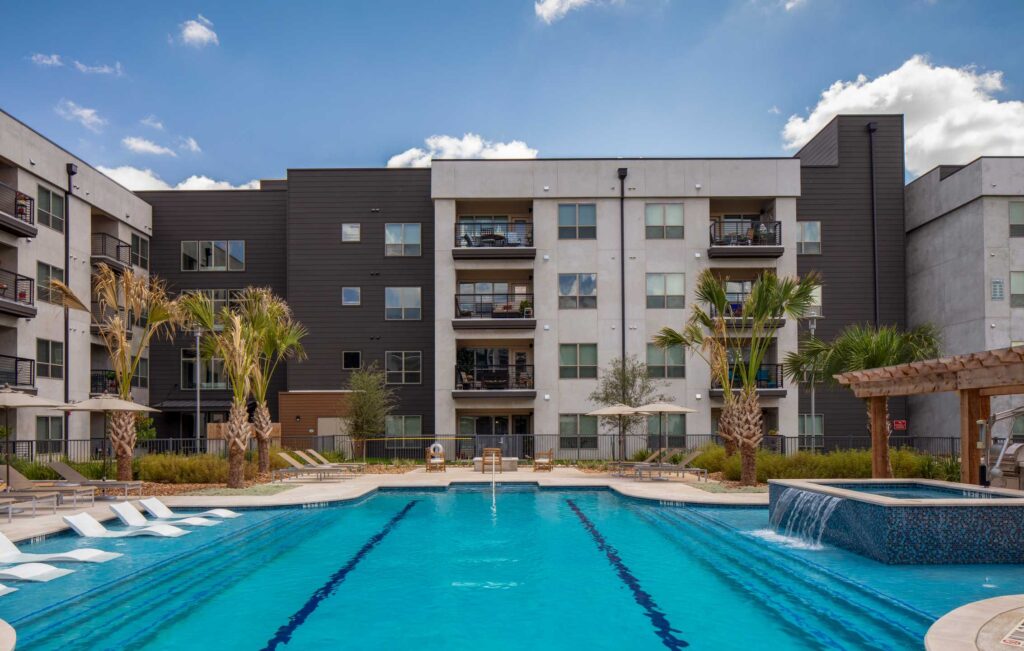 Modern apartment complex featuring a vibrant pool area, lounge chairs, and palm trees, set against a clear blue sky.