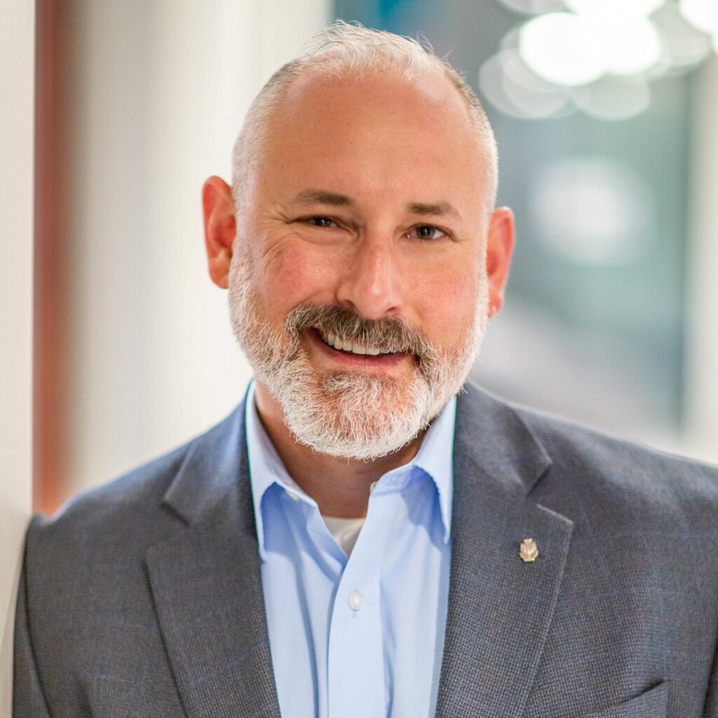 A smiling man with a greying beard is dressed in a blazer and light blue shirt, standing against a softly blurred background.