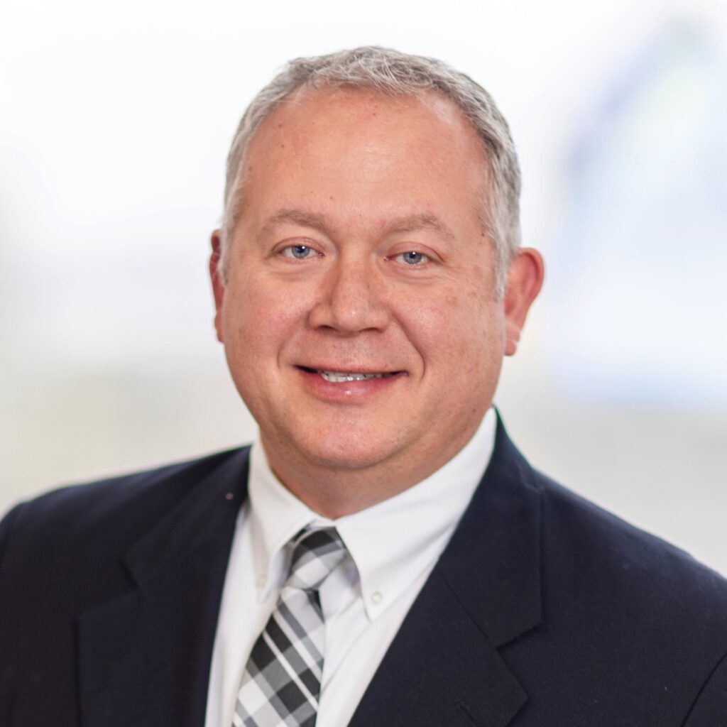 A man in a suit and tie smiles confidently, showcasing a professional appearance with a neutral background.