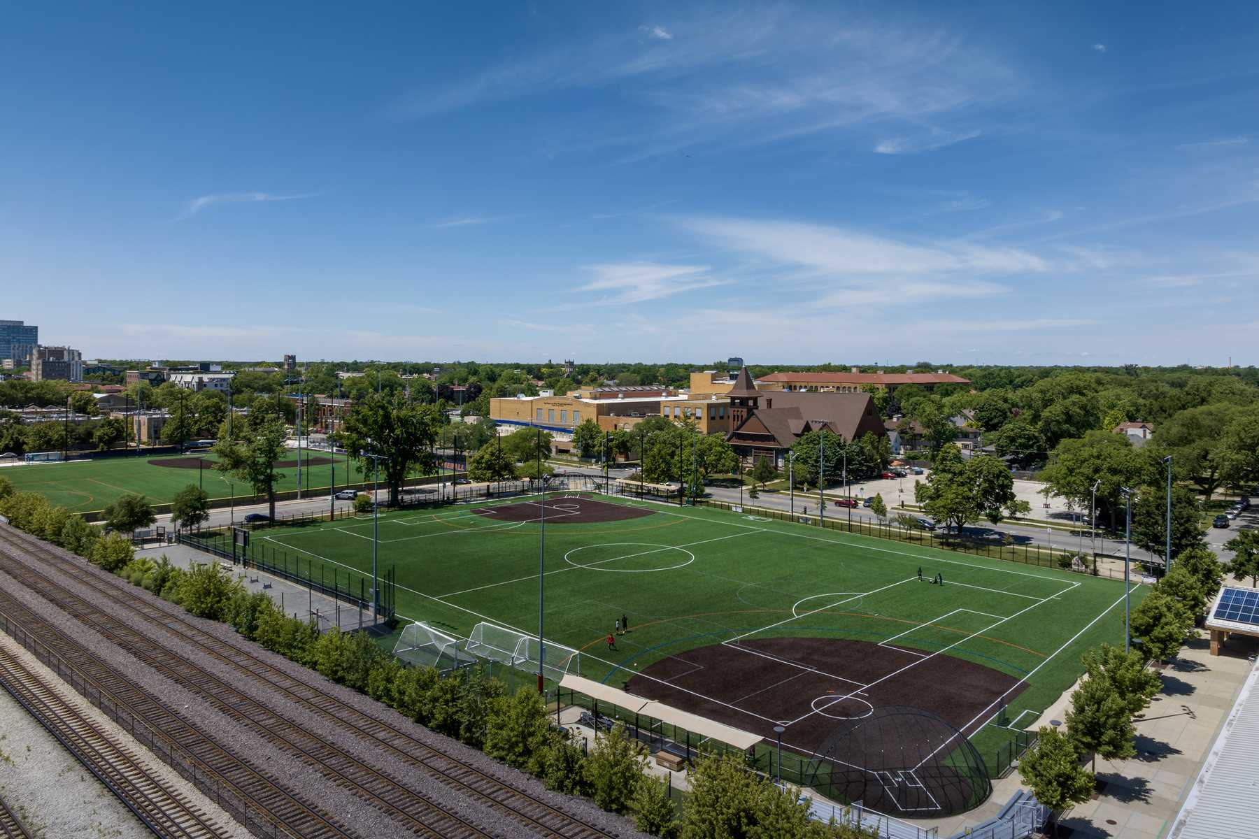 A sunlit urban park features multiple soccer fields, surrounded by trees and buildings, with a clear blue sky overhead.