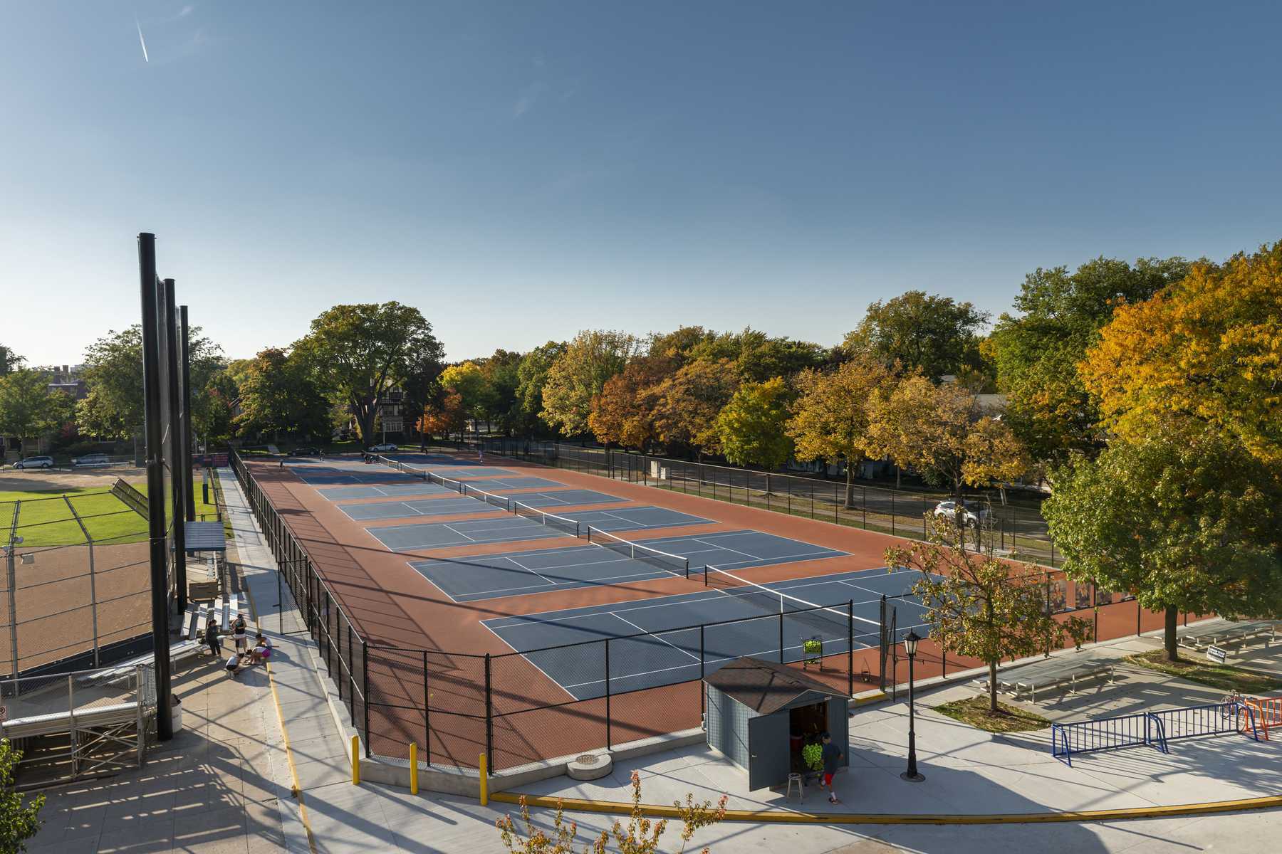 A spacious outdoor tennis complex features several courts amidst vibrant autumn foliage under a clear blue sky. Activity fills the area.