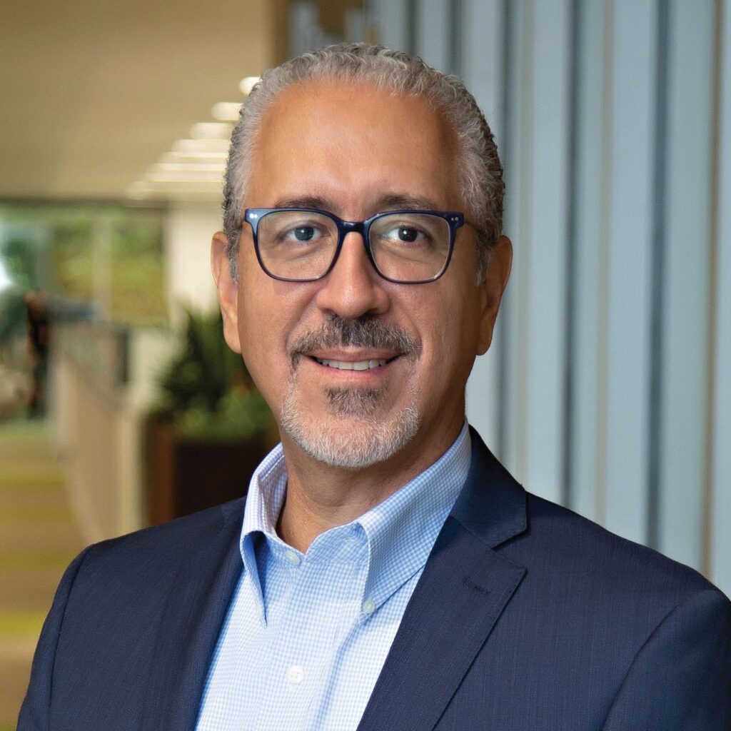 A professionally dressed man with glasses smiles confidently in a modern office setting, featuring natural light and greenery in the background.