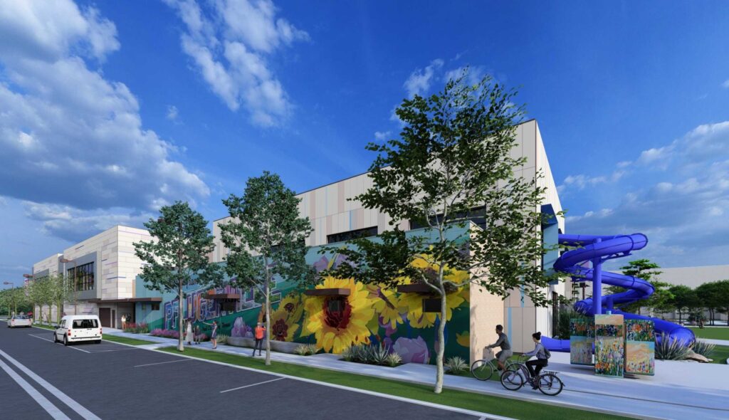 A modern building features colorful murals, green landscaping, and a vibrant blue water slide, set against a bright, cloudy sky.