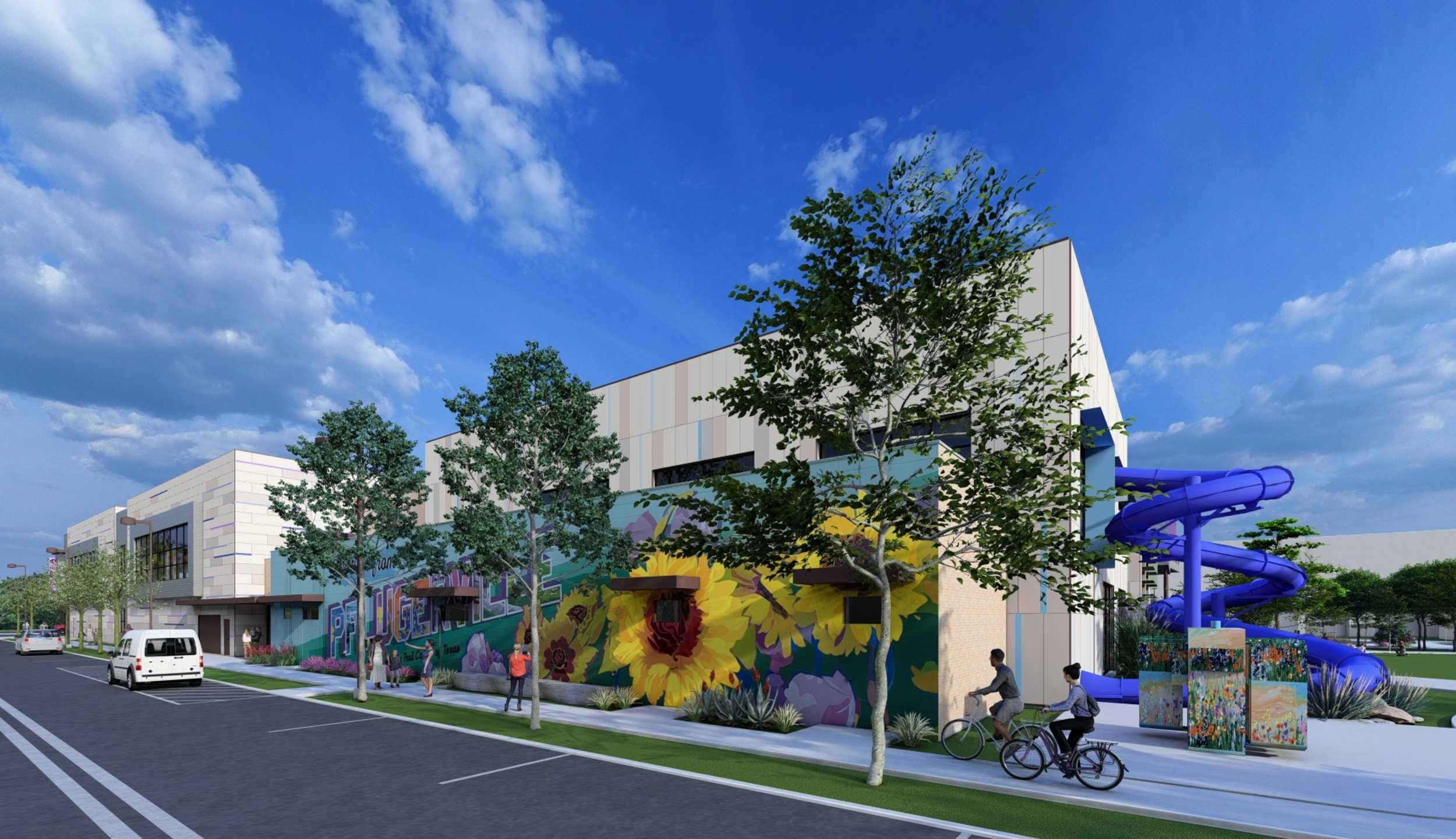 A modern building features colorful murals, green landscaping, and a vibrant blue water slide, set against a bright, cloudy sky.