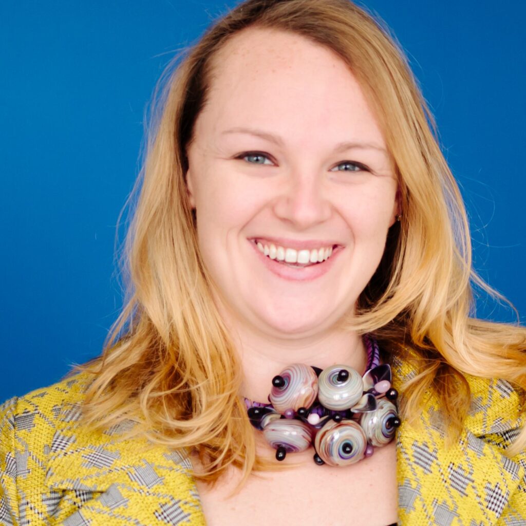 A smiling person with long, wavy hair wears a vibrant yellow patterned blazer and a colorful, artistic necklace against a blue background.