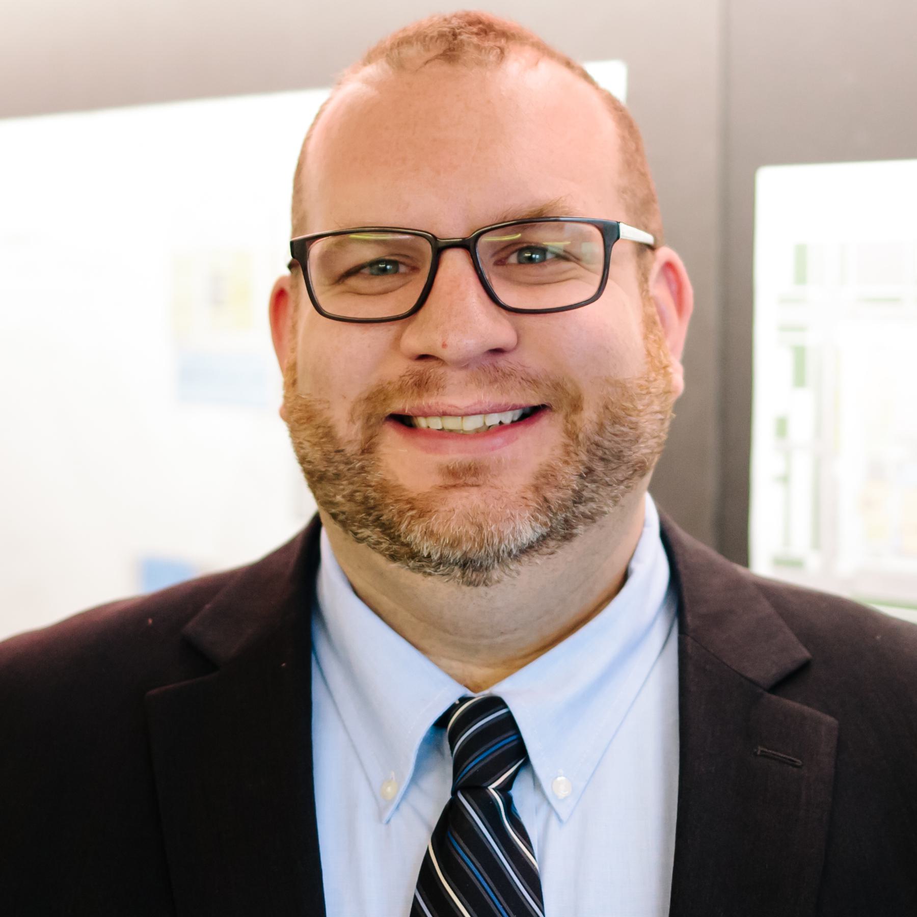 A smiling man with glasses and a beard wears a black suit and a light blue shirt, exuding confidence and approachability in a professional setting.