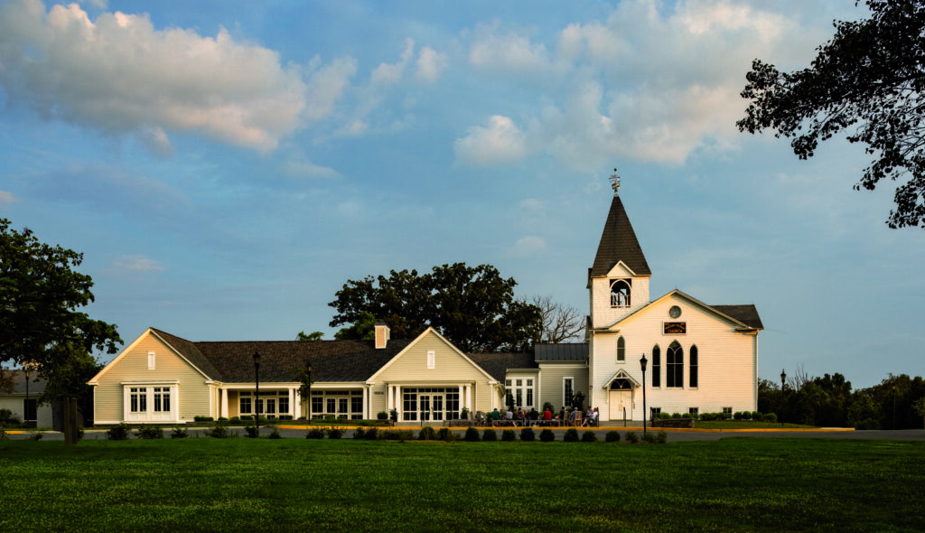 A charming white church with a tall steeple, accompanied by a modern building, set against a backdrop of blue skies and greenery.