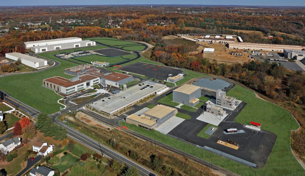 An aerial view of a complex featuring several modern buildings, parking areas, and landscaped green spaces, set against a backdrop of autumn foliage.