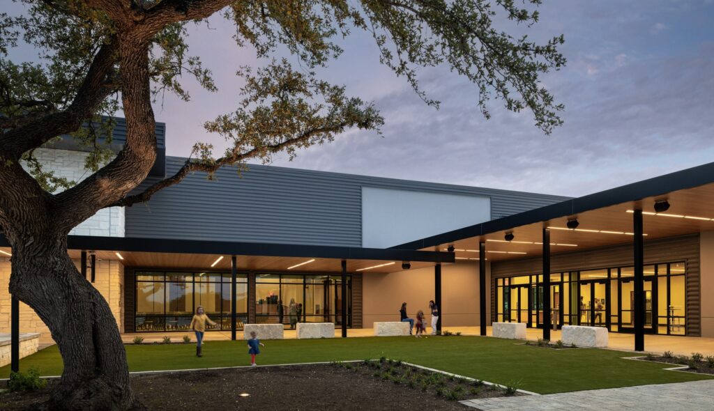 A modern building features large glass windows, outdoor seating, and a landscaped area with children playing under a mature tree at dusk.
