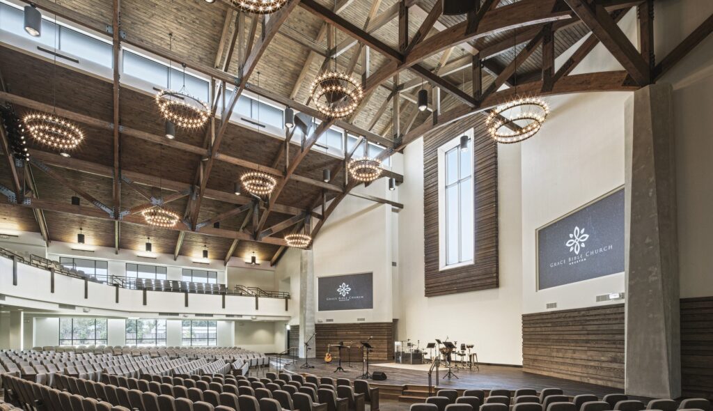 A spacious, modern auditorium features wooden beams, chandeliers, and rows of seating, with screens displaying a logo at the front.