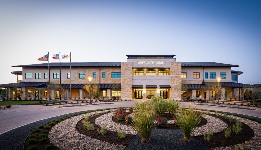 A modern building featuring stone accents and large windows, surrounded by landscaped gardens and flags, creating an inviting entrance.