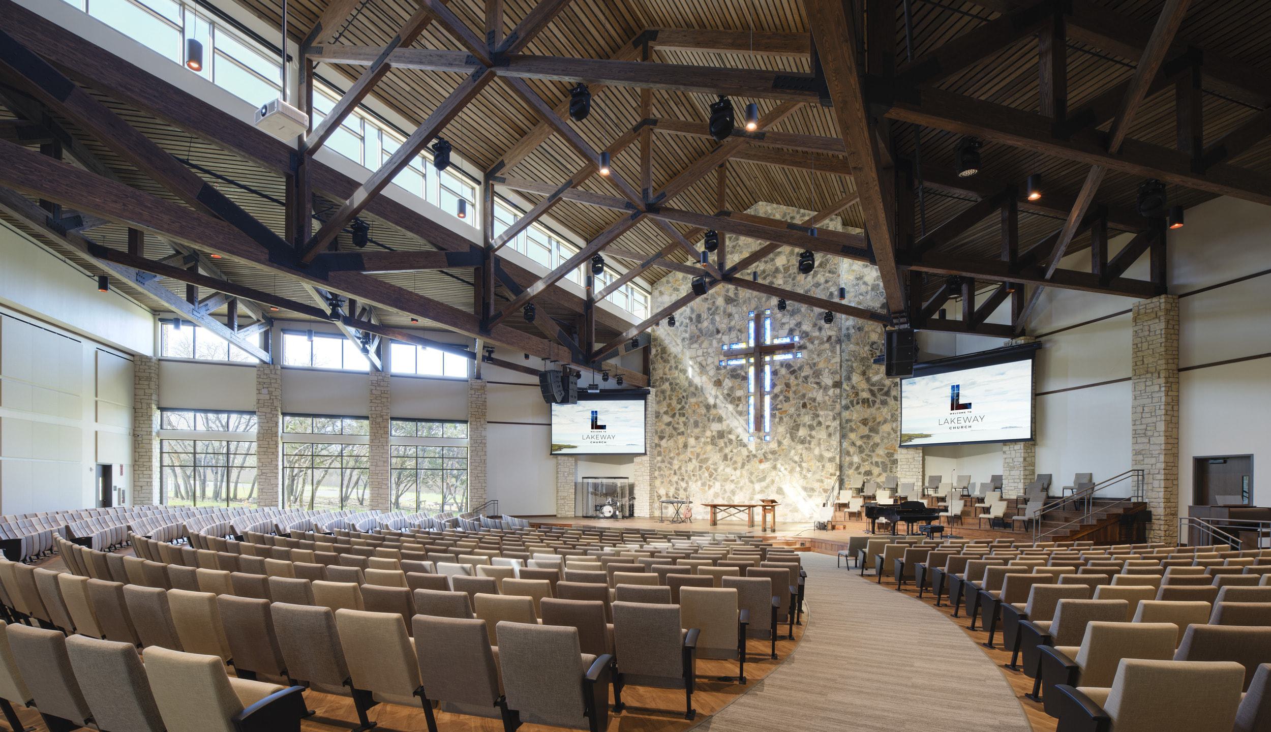 A spacious, modern worship hall featuring a wooden ceiling, a prominent cross, rows of seats, and large screens for presentations.