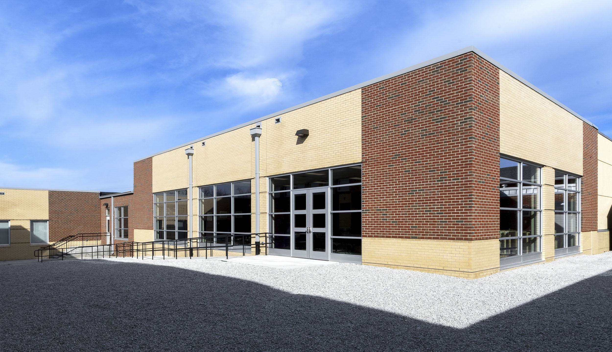A modern building features a mix of brick and glass walls with a ramp leading to the entrance, set against a bright blue sky.