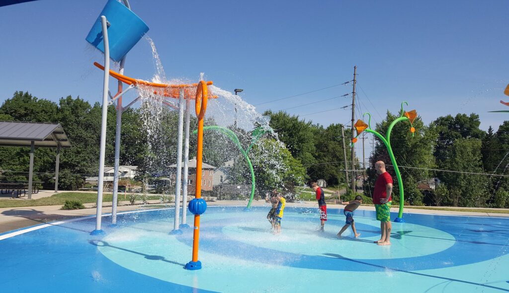 Children play joyfully in a splash pad, enjoying water features under a clear blue sky, while an adult supervises nearby.