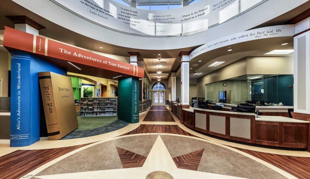 A vibrant library interior features oversized book decorations, inviting spaces, and an open layout with wood and carpet flooring.