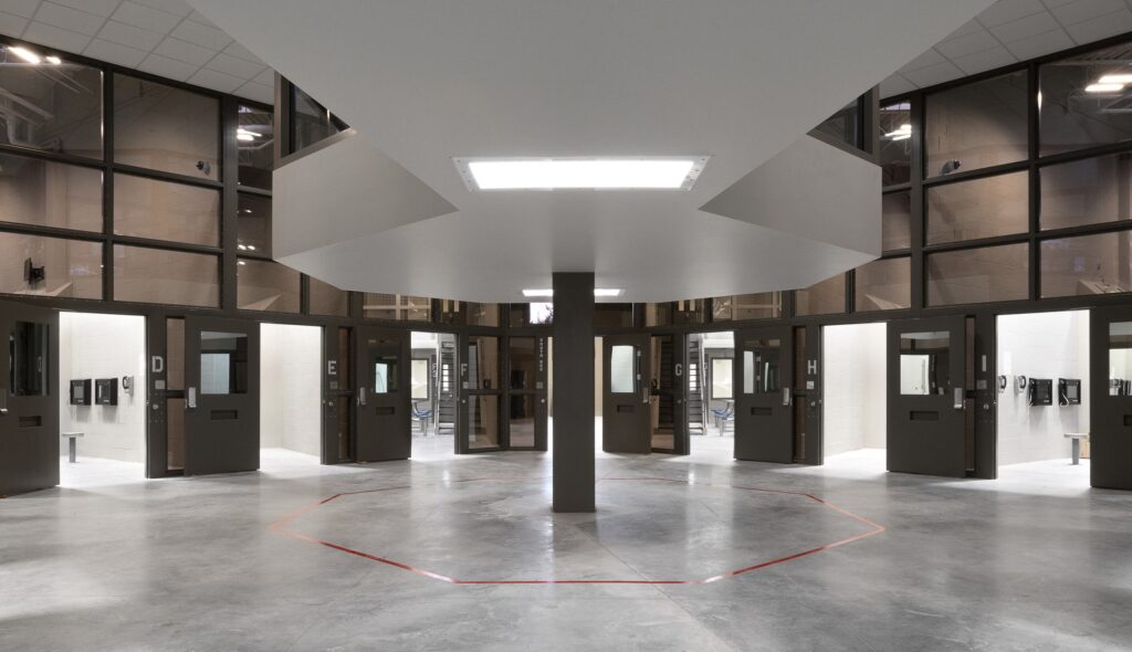 A modern, circular prison corridor with multiple closed doors, open to a central area illuminated by overhead lighting and surrounded by glass walls.