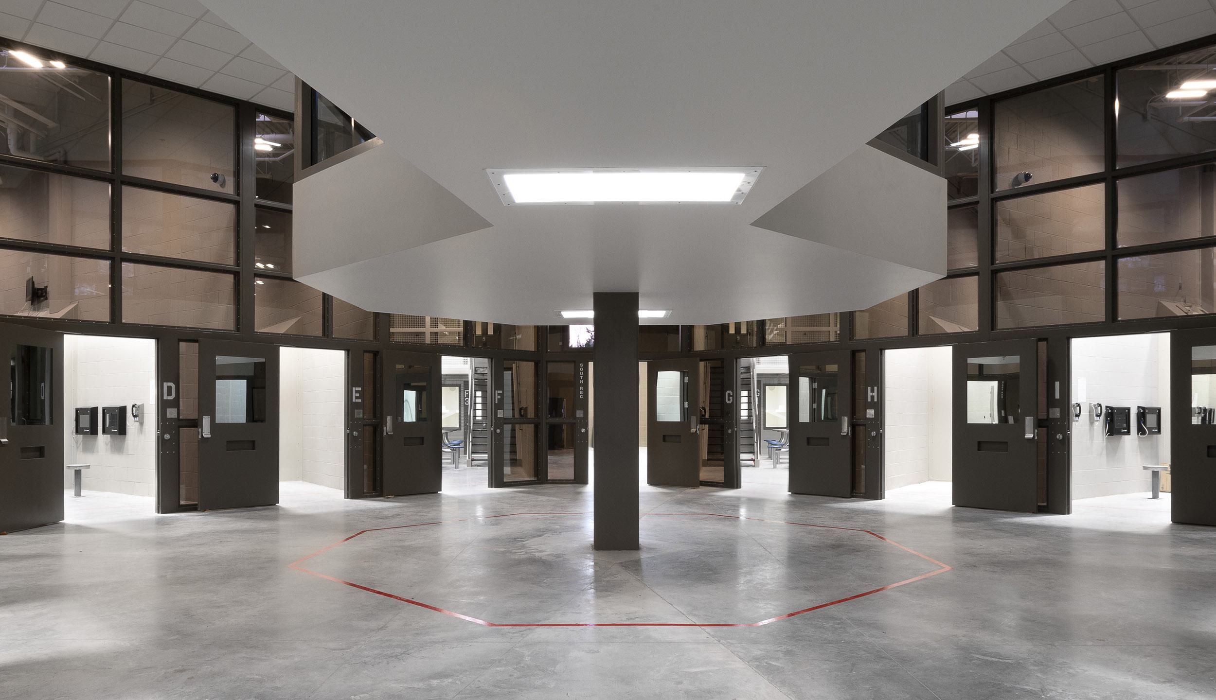 A modern, circular prison corridor with multiple closed doors, open to a central area illuminated by overhead lighting and surrounded by glass walls.