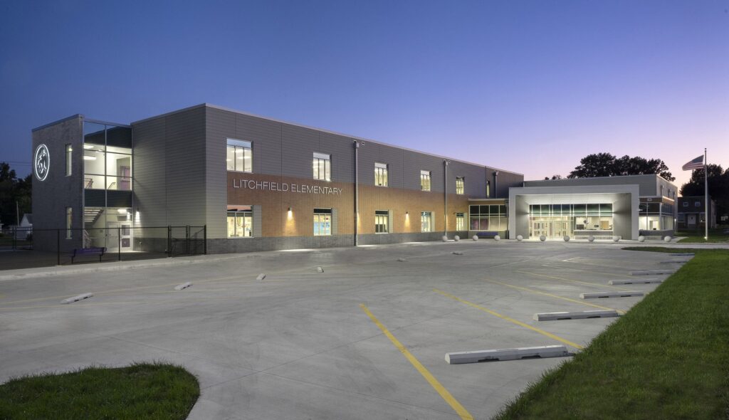 A modern, well-lit elementary school building features large windows and a spacious outdoor area, set against a twilight sky.