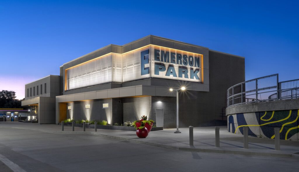 A modern building features prominent "EMERSON PARK" signage, well-lit with a sleek design, surrounded by landscaped areas and a vibrant mural.