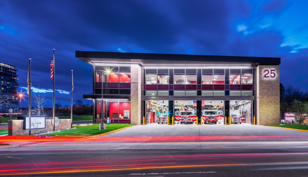 Modern fire station with illuminated bays, large windows, and a sleek design. Flags stand outside against a darkening sky.