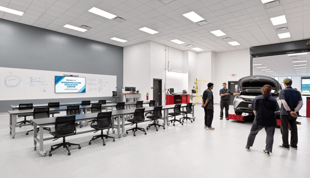 A modern automotive training facility featuring a classroom area and a car with its hood open, with instructors and students engaged in learning.