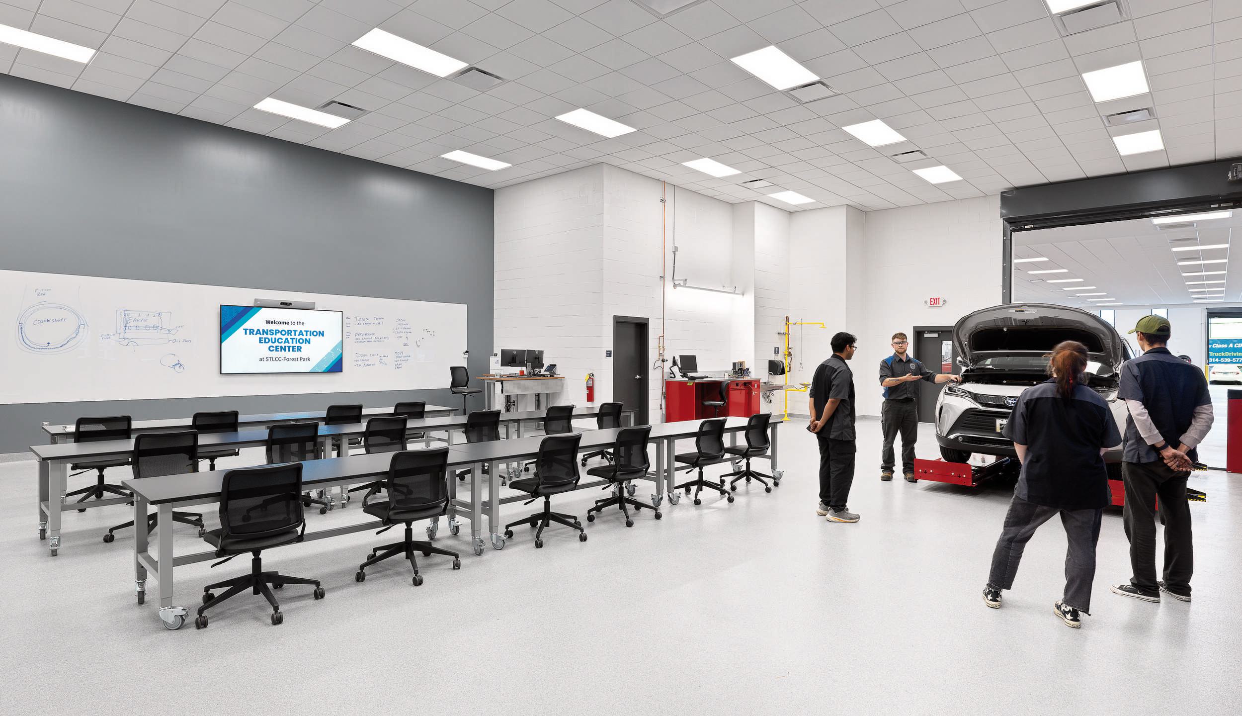 A modern automotive training facility featuring a classroom area and a car with its hood open, with instructors and students engaged in learning.